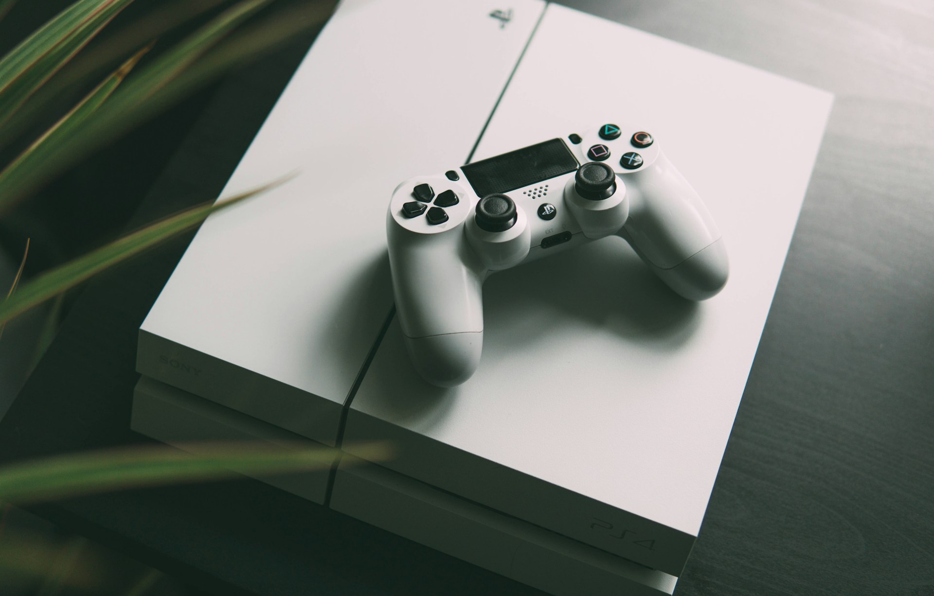 A white gaming console is placed on a dark surface, with a matching white controller resting on top. The console features smooth, clean lines and a sleek design. The surrounding area is slightly blurred, giving focus to the gaming equipment.