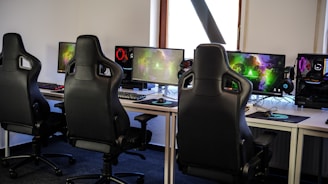 A row of gaming stations equipped with large monitors, gaming chairs, keyboards, and headphones. The setup is organized and ready for gaming, featuring a colorful screen display. The room has a modern and tech-savvy ambiance.