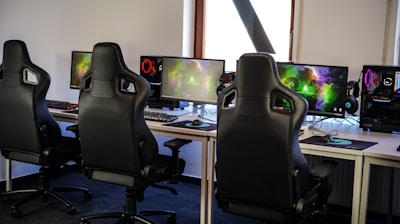 A row of gaming stations equipped with large monitors, gaming chairs, keyboards, and headphones. The setup is organized and ready for gaming, featuring a colorful screen display. The room has a modern and tech-savvy ambiance.