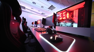 A row of high-end gaming computers on sleek desks, featuring RGB lighting and gaming accessories. The computers are housed in transparent cases with visible components, emitting a red glow. Comfortable gaming chairs with ergonomic design are positioned next to the desks, and the room's ceiling is modern with recessed lighting.