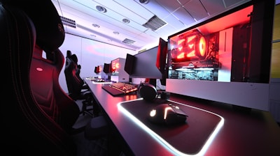 A row of high-end gaming computers on sleek desks, featuring RGB lighting and gaming accessories. The computers are housed in transparent cases with visible components, emitting a red glow. Comfortable gaming chairs with ergonomic design are positioned next to the desks, and the room's ceiling is modern with recessed lighting.