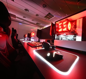A modern gaming setup featuring several high-end computers with illuminated red interior lighting. The room is equipped with gaming chairs exhibiting a sleek black and red design. Multiple monitors are arranged on a long desk, and peripheral devices like keyboards and mice are uniformly placed, reflecting an ambiance ideal for gaming or competitive play.