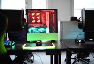 A modern gaming setup is displayed on a desk with multiple computer monitors and an open gaming PC tower. The tower is illuminated with red and green LED lights, showcasing a 'GeForce RTX' graphics card. A gaming mouse rests on a mousepad with glowing edges, and there is a keyboard with vibrant backlighting. The room appears well-lit with visible windows in the background.