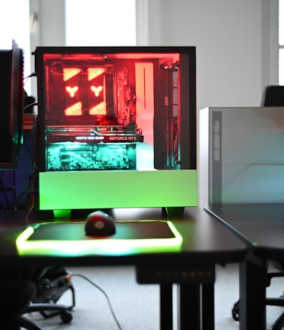 A modern gaming setup is displayed on a desk with multiple computer monitors and an open gaming PC tower. The tower is illuminated with red and green LED lights, showcasing a 'GeForce RTX' graphics card. A gaming mouse rests on a mousepad with glowing edges, and there is a keyboard with vibrant backlighting. The room appears well-lit with visible windows in the background.