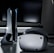 A sleek virtual reality headset sits on a reflective surface beside a gaming console and its controller. The background features a large dark monitor screen, creating a modern and futuristic setting. The lighting is subdued, highlighting the glossy surfaces of the electronic devices.