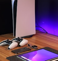 A sleek, modern setup featuring a gaming console with two controllers on a wooden surface. Nearby are a smartphone, smartwatch, tablet with a vivid screen, wireless earbuds, and a stylus. The background is illuminated with a soft purple glow.