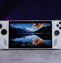 A handheld gaming console with a central screen displaying a scenic wallpaper of mountains and a lake, flanked by controller buttons and joysticks on both sides. The lighting around is dim, with purplish hues in the background, creating a modern and elegant atmosphere.