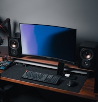 A modern gaming and streaming setup with a curved monitor placed centrally on a wooden desk. Surrounding the monitor are powerful speakers and a high-end PC tower with glowing fans. Various peripherals, including a mechanical keyboard, ergonomic mouse, and audio mixer, are neatly arranged. Overhead, a professional microphone and stand are ready for use, possibly for gaming commentary or podcasting.