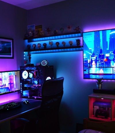 A vibrant gaming setup featuring multiple monitors displaying a cyberpunk cityscape. The room is illuminated with neon purple and blue LED lights, adding a futuristic ambiance. A custom-built PC with glowing components sits to the side, and shelves with collectible figurines are visible. A large digital screen on the wall shows an animated city scene, enhancing the immersive atmosphere.